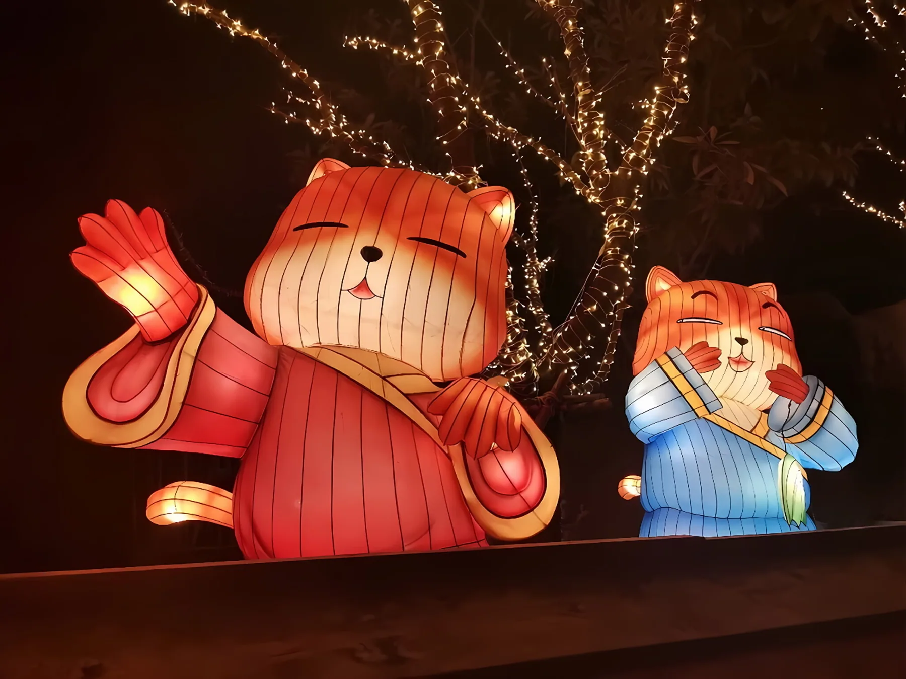 Waving Miss Cat lantern characters under tree lights at night